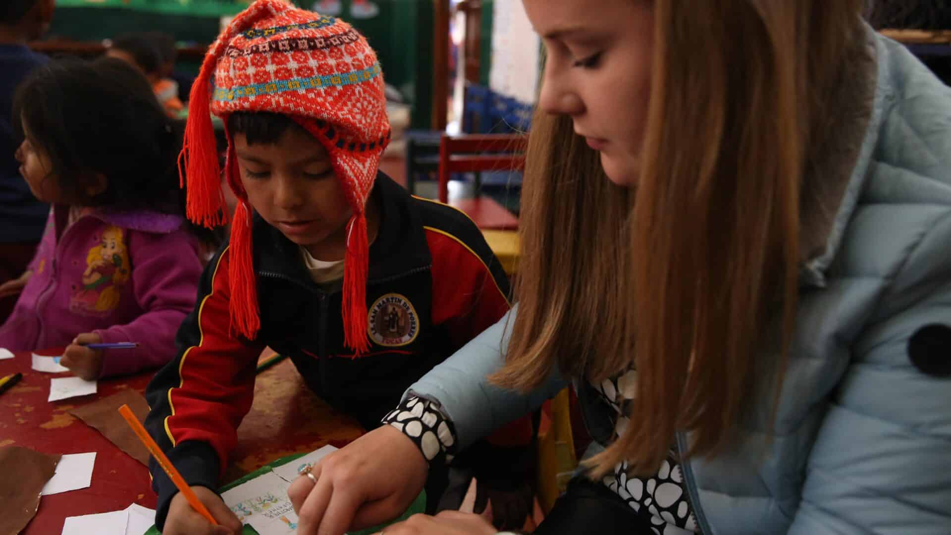 A volunteer working with children in Peru sits with a child in a classroom to help them with an activity.