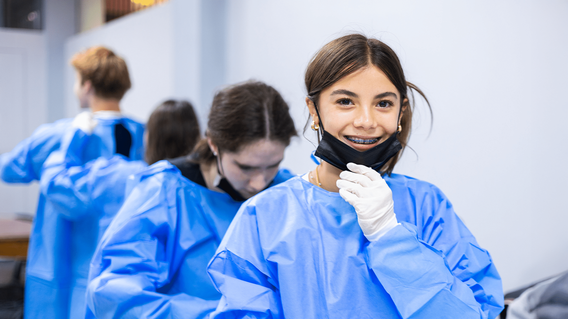 A teenage girl with braces and a bright smile looks at the camera while adjusting a black face mask under her chin. She is wearing blue medical scrubs/gowns and white protective gloves. In the softly blurred background, other teenagers are also putting on matching blue medical scrubs in a brightly lit room.