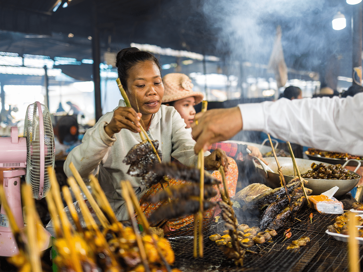 A woman prepares and grills various foods on long wooden skewers at a bustling indoor market stall. Smoke rises from the large grill filled with fish and local delicacies, while a pink electric fan sits to the left and a customer reaches in from the right.