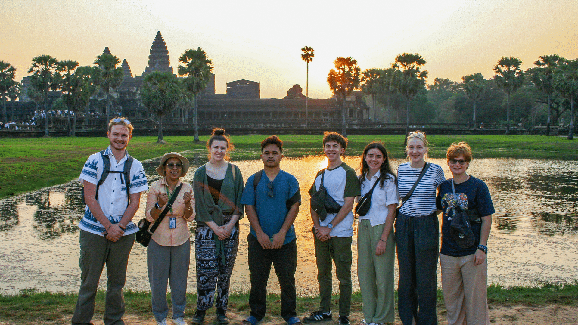 A group of eight people stand in a line smiling in front of a reflective pond at sunrise or sunset. In the background, the distinctive towers of the ancient Angkor Wat temple complex in Cambodia are silhouetted against a golden sky with palm trees.