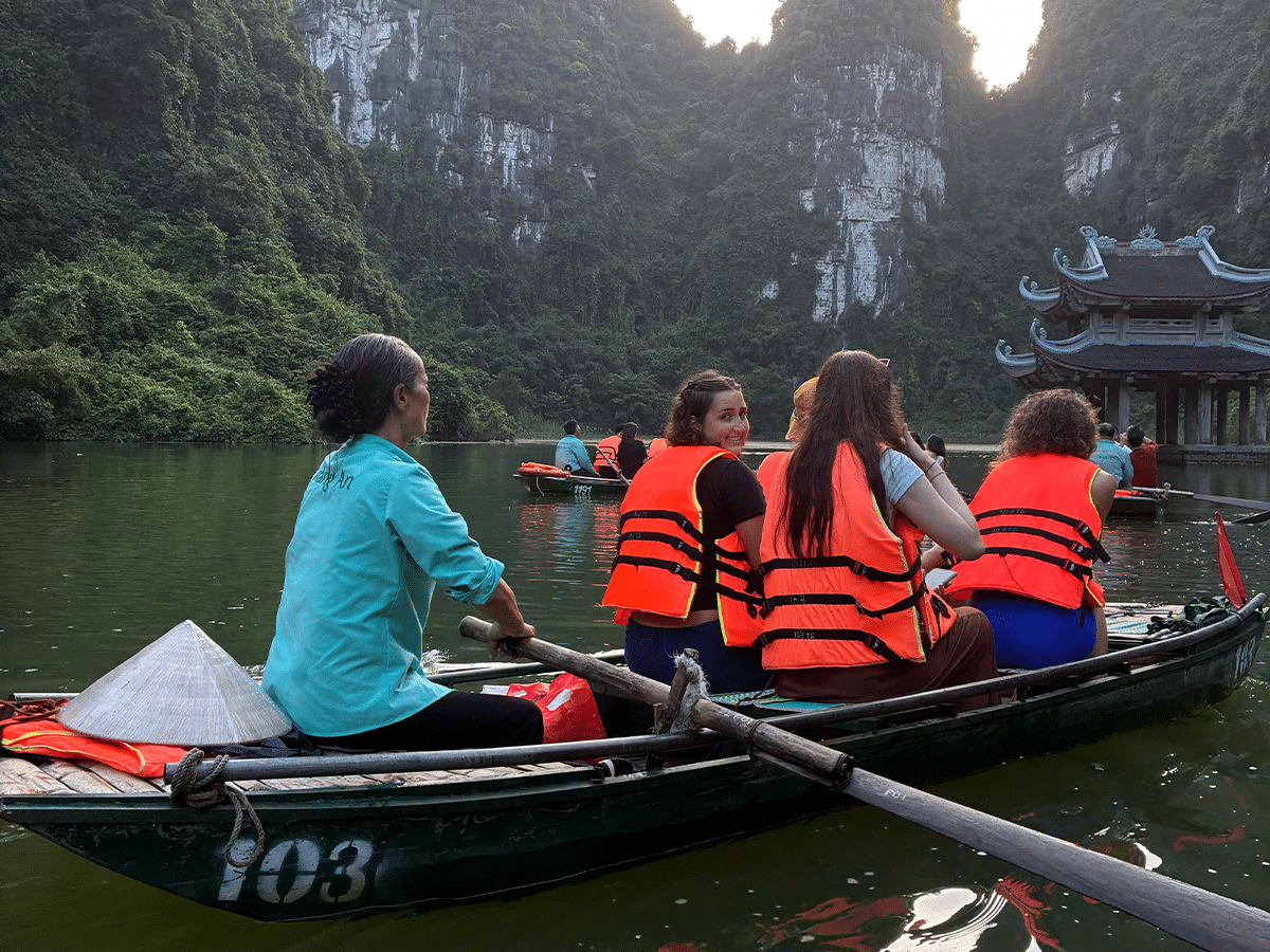 Three young women wearing bright orange life jackets sit in a small rowing boat steered by a local guide in a light blue shirt. They are traveling down a river in Trang An, Vietnam, surrounded by towering, green limestone cliffs and other tourist boats near a traditional pavilion.
