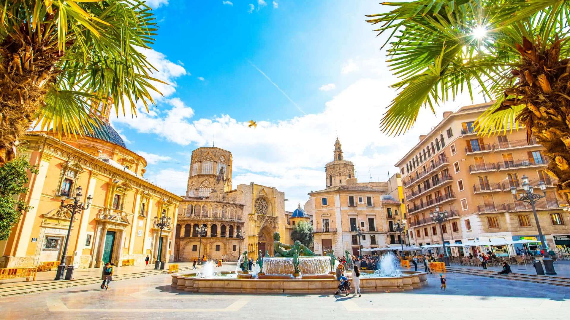 A sunlit view of Plaza de la Virgen in Valencia, Spain, framed by palm trees. A large fountain sits in the center of the stone square, surrounded by historic buildings, including the Valencia Cathedral and Basilica under a bright blue sky.