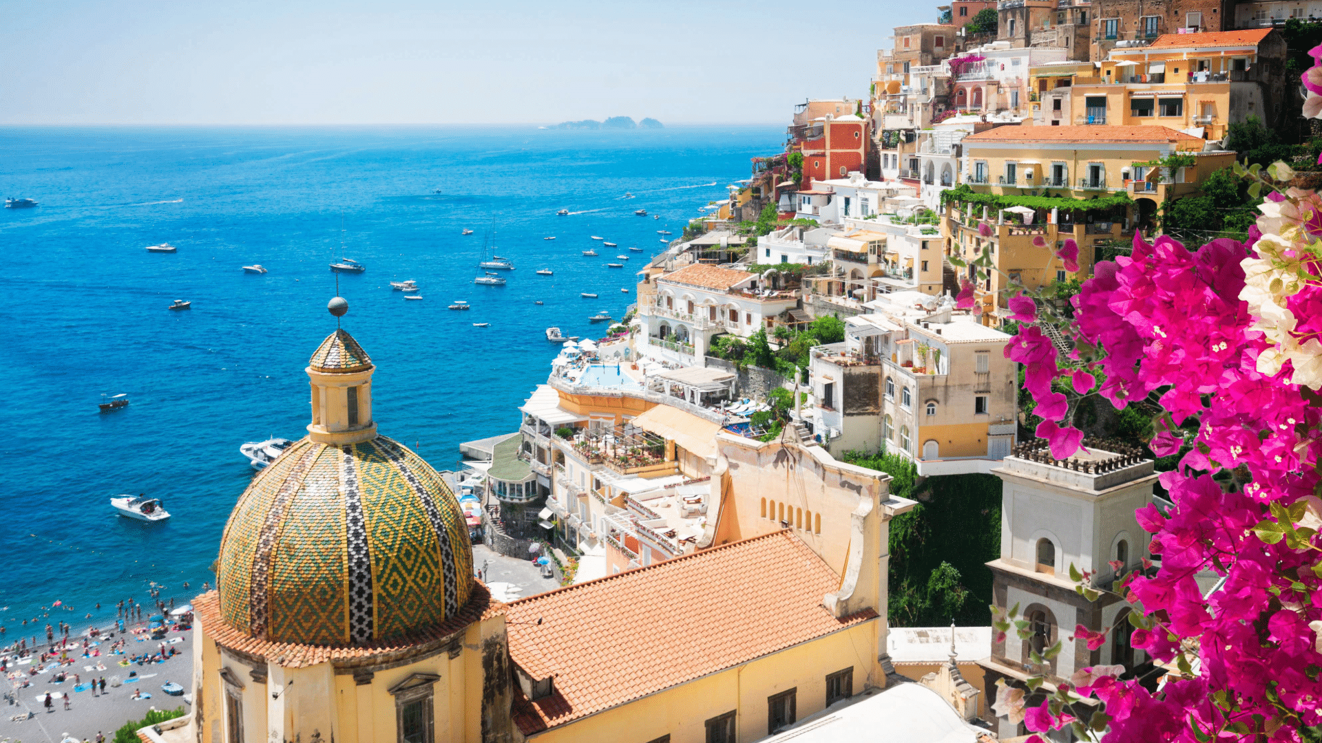 An elevated landscape photograph from Italy. In the right foreground, bright magenta bougainvillea blossoms frame the view. Below in the left foreground stands the tiled dome of the Church of Santa Maria Assunta, decorated in yellow, green, and blue geometric majolica patterns. Warm pastel-coloured buildings cling to the steep cliffside, cascading down to a beach filled with umbrellas and people. The deep blue sea stretches out to the horizon, dotted with small boats and yachts under a clear blue sky.