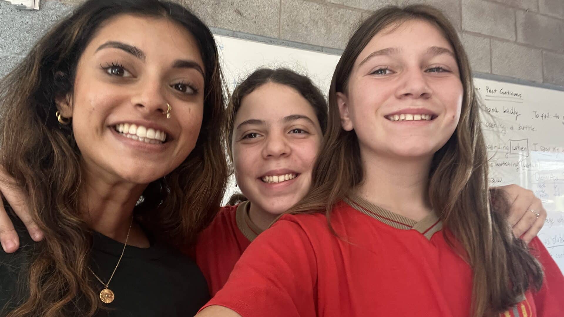 A cheerful group selfie of a young woman with long dark hair and two young girls wearing matching red shirts, all smiling together inside a classroom setting.