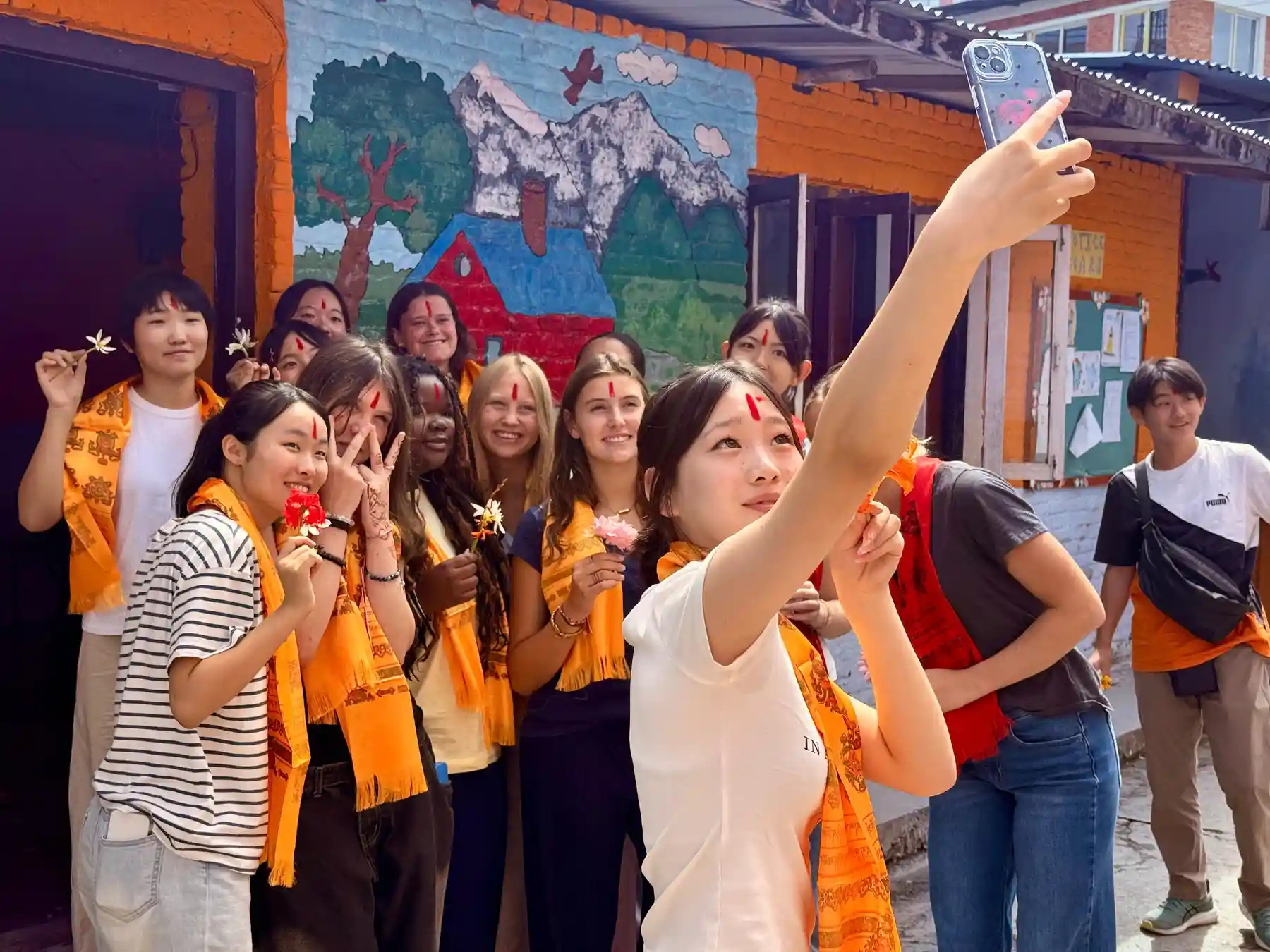 Group of young travelers wearing traditional scarves and tika marks taking a selfie in front of a painted mural.